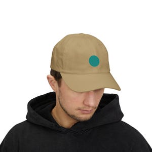 May include: A tan baseball cap featuring a solid teal circle centered on the front panel. The cap has a structured crown, a curved brim, and a series of stitched eyelets on the top panels. The design is simple, clean, and minimalist in its presentation.