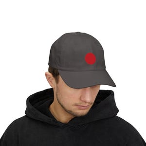 May include: A charcoal gray baseball cap featuring a solid, circular red graphic centered on the front panel. The cap has a structured crown, a curved brim, and a matching top button, providing a clean and simple design for everyday headwear.
