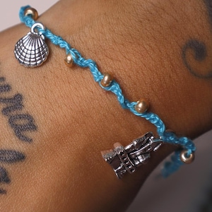 Puede incluir: Una pulsera de cordón trenzado azul claro con pequeñas cuentas metálicas de tono dorado intercaladas a lo largo. La pulsera incluye dos dijes de tono plateado, uno con forma de concha marina y otro con forma de faro.
