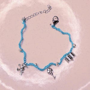 May include: A light blue cord bracelet featuring three silver-toned metal charms: a rabbit, a key with a floral head, and an open book. The bracelet includes small silver-toned beads interspersed along the cord, a lobster claw clasp, and an extender chain.