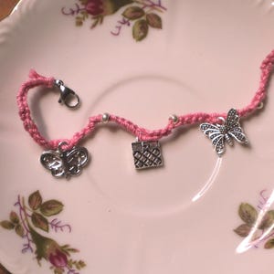 May include: A pink braided cord bracelet featuring three silver-toned metal charms: two butterflies and one square gift box. The bracelet includes small silver-toned beads interspersed along the cord and a silver-toned lobster claw clasp closure.
