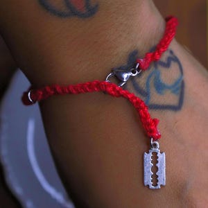 Op de afbeelding: Een rode gevlochten koordarmband met een zilverkleurige metalen scheermesje bedel en een karabijnsluiting. De armband is gemaakt van dun, getextureerd rood touw en bevat een kleine metalen ring bij het sluitingsmechanisme.