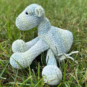 May include: A blue and green plush toy animal with a long tail and four legs. The toy is sitting on a bed of green grass.