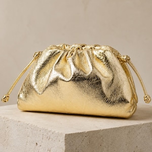 Puede incluir: Un bolso de mano dorado metalizado con un diseño fruncido en la parte superior. El bolso cuenta con un cierre de cordón en ambos lados con finos cordones dorados que terminan en pequeños nudos, creando una forma redondeada y una textura brillante.