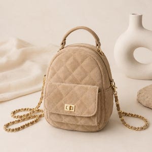 May include: A beige quilted mini backpack featuring a soft-touch texture, a top handle, and a front pocket with a gold-tone metal turn-lock closure. The bag includes thin gold-tone chain straps interwoven with matching beige material for carrying.