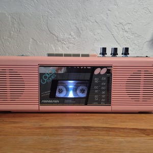 May include: A pink Soundesign Color Tunes portable cassette player and radio featuring a central cassette deck, a radio frequency display, three black control knobs on the top, and integrated speakers on the left and right sides.