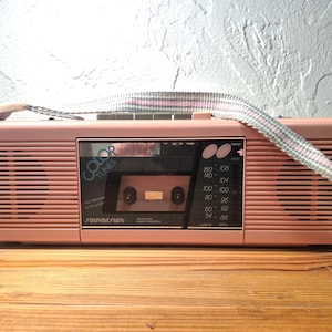 May include: A pastel pink portable cassette player and radio with a horizontal ribbed design. The front panel features the text Color Tunes and Soundesign, a cassette deck, and a radio frequency tuner dial. A woven carrying strap is attached.