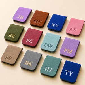 May include: A collection of twelve rectangular felt magnetic bookmarks in various colors including purple, brown, blue, pink, olive green, red, teal, tan, gray, and dark blue. Each bookmark features two capital letters embossed in gold or white.