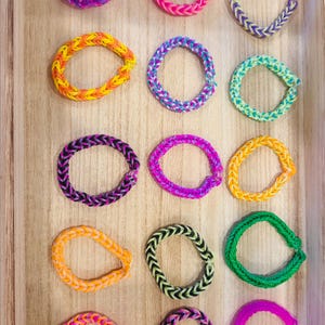 May include: Fifteen circular bracelets made of small, interlocking rubber bands in various colors, including purple, pink, orange, yellow, green, black, and multi-colored patterns, arranged in a grid of five rows and three columns.