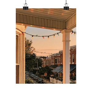 May include: A digital art print depicting a view from a porch with a white column, looking out over a street at dusk. A string of colorful holiday lights hangs across the frame. Below, a residential street features parked cars and brick houses.