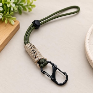 Puede incluir: Un cord&oacute;n de paracord verde oliva con un cierre de pl&aacute;stico negro, una secci&oacute;n envuelta en cuerda con patr&oacute;n beige y negro, y un mosquet&oacute;n de metal negro unido al extremo para asegurar objetos peque&ntilde;os o accesorios.
