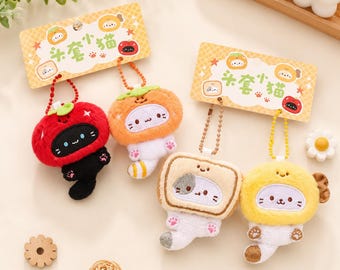 May include: Four plush cat keychains attached to two yellow rectangular display cards. The cats are dressed in fuzzy costumes resembling a red tomato, an orange, a slice of toast, and a yellow lemon, each featuring a small metal ball chain.