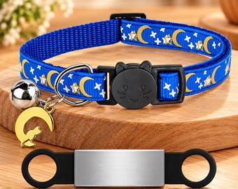 May include: A blue pet collar featuring a yellow crescent moon and white star pattern, a black cat-shaped plastic buckle, a silver bell, and a gold crescent moon charm. Below the collar is a rectangular silver metal tag with black side loops.