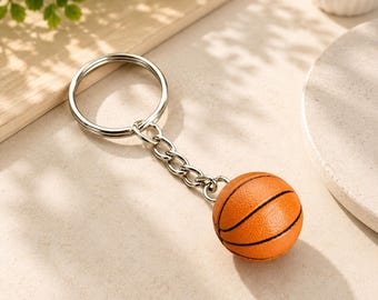 May include: A basketball keychain featuring a small, textured orange sphere with black lines attached to a silver-toned metal split ring by a short, sturdy metal chain. The basketball charm has a realistic pebbled surface and classic design.
