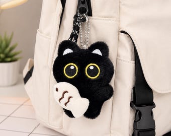 May include: A plush black cat keychain with large yellow eyes and white inner ears, holding a small white fish toy. The cat is attached to a cream-colored backpack by a silver ball chain and a black and white patterned cord, featuring soft fur.