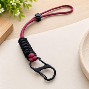 Puede incluir: Un cord&oacute;n con una cuerda granate con un regulador de pl&aacute;stico negro, una secci&oacute;n de paracord negro y granate tejido en un patr&oacute;n de punto cobra, y un mosquet&oacute;n de metal negro sujeto al extremo de la secci&oacute;n de cuerda trenzada.