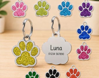 May include: A collection of paw-shaped pet ID tags in various glittery colors including pink, lime green, blue, purple, yellow, green, black, and orange. One silver-toned tag features the engraved name Luna and the number 01234 567890.