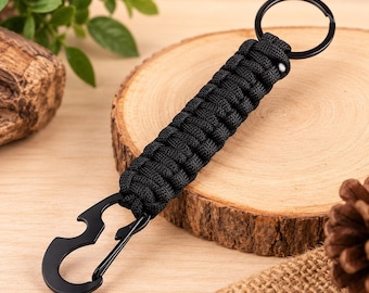 May include: A black paracord keychain featuring a braided nylon cord handle, a circular metal split ring at the top, and a matte black metal carabiner with an integrated bottle opener at the bottom, providing a functional tool for everyday carry.