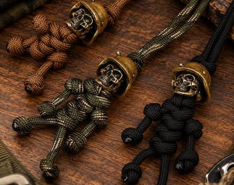 May include: Three paracord keychains shaped like figures, each featuring a brass-colored metal skull wearing a helmet. The figures are crafted from brown, olive green, and black woven cord, with knotted limbs and a loop at the top for attachment.