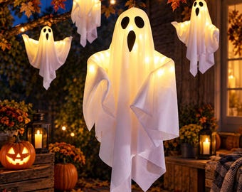 May include: Four white hanging ghost decorations made of fabric, each featuring black oval eyes and a mouth. These illuminated figures are suspended from tree branches, glowing with warm internal lights, set against an outdoor autumn scene.