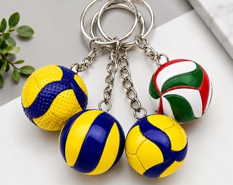 May include: A set of four spherical volleyball keychains attached to silver metal rings and chains. The keychains feature various color combinations including yellow and blue, as well as a white, green, and red design, all with textured surfaces.