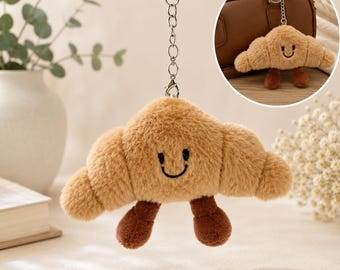 May include: A plush croissant keychain featuring a light brown fuzzy texture, a stitched black smiling face, and two small dark brown dangling legs. The item is attached to a silver-toned metal chain and lobster clasp for secure hanging.
