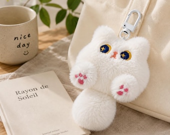 May include: A white plush cat keychain featuring embroidered dark blue eyes with yellow crescent moon details, a small pink nose, and pink paw pads on the front feet. The cat is attached to a white metal lobster clasp for easy hanging.