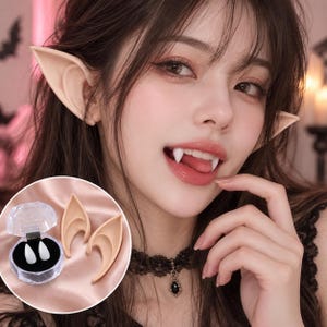 May include: A set of beige pointed elf ears and two white vampire fangs. The fangs are displayed inside a small, clear, diamond-shaped plastic case with a black interior lining, while the two ear pieces are positioned next to the container.