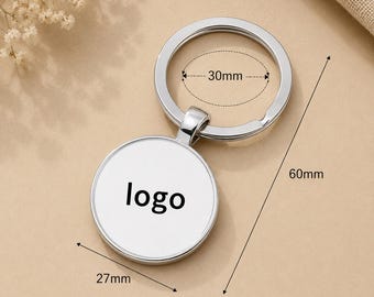 May include: A silver-colored circular keychain with a white center insert featuring the black text logo. The keychain has a total length of 60mm, a circular pendant diameter of 27mm, and a split ring inner diameter of 30mm as indicated by lines.