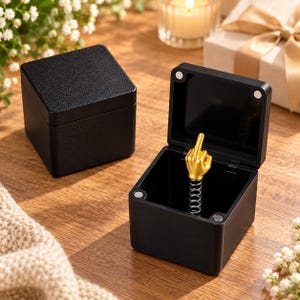 May include: Two square black boxes with textured lids. One box is closed, while the other is open, revealing a gold-colored hand figurine making an obscene gesture mounted on a silver metal spring inside the center of the box base.