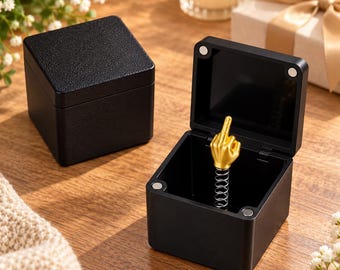 Puede incluir: Dos cajas cuadradas negras con tapas texturizadas. Una caja está cerrada, mientras que la otra está abierta, revelando una figura de mano dorada que hace un gesto obsceno montada sobre un resorte de metal plateado en el centro de la base.