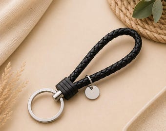 May include: A black braided leather keychain featuring a silver-toned metal ring and a small, circular, blank silver-toned metal charm attached to the base. The braided loop is secured by a black cylindrical connector piece made of metal.