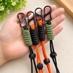 Puede incluir: Cuatro cordones con mosquetones negros unidos a secciones de paracord trenzado. Dos cordones tienen un patr&oacute;n de color verde ne&oacute;n y negro, mientras que dos son de color negro y naranja s&oacute;lido. Cada uno incluye cierres de pl&aacute;stico negro.