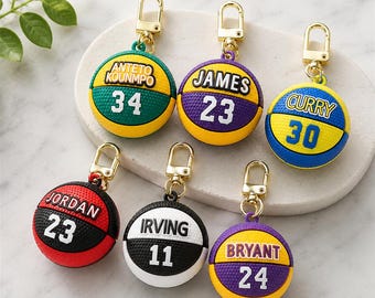 May include: Six basketball-shaped keychains with gold-tone lobster clasps. Each features a player name and number: Antetokounmpo 34, James 23, Curry 30, Jordan 23, Irving 11, and Bryant 24. The balls are textured and come in various color combinations.
