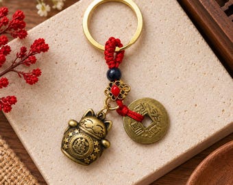 May include: A gold-toned metal keychain featuring a small beckoning cat charm and a round coin-shaped pendant with a square center hole. The charms are attached to a gold ring by a red knotted cord with a dark blue bead and a small red accent.