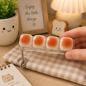 May include: A clear plastic keychain featuring four square, white, and orange translucent charms that resemble small, rounded, glossy fried eggs. The keychain includes a silver-toned metal ring and a short chain link attached to one end of the clear rectangular base.