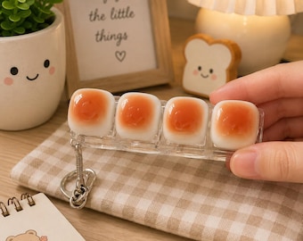 May include: A clear plastic keychain featuring four square, white, and orange translucent charms that resemble small, rounded, glossy fried eggs. The keychain includes a silver-toned metal ring and a short chain link attached to one end of the clear rectangular base.