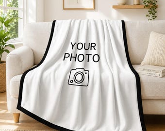 May include: A white throw blanket with a black border featuring the black text "YOUR PHOTO" and a simple black line-art icon of a camera centered on the fabric. The blanket is draped over a light-colored sofa, displaying a smooth texture.