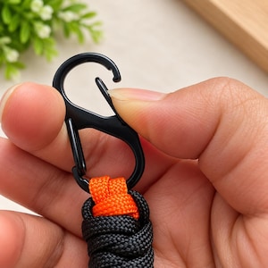 Puede incluir: Un mosquet&oacute;n de metal negro unido a un llavero trenzado. El llavero presenta un tejido grueso y texturizado hecho de cuerda negra con un acento de cuerda de color naranja brillante en la parte superior donde se conecta al anillo met&aacute;lico.