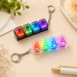 Puede incluir: Dos llaveros de interruptores de teclado mecánico, cada uno con cuatro teclas cuadradas y translúcidas en fila. El llavero superior tiene bases negras, mientras que el inferior tiene bases transparentes. Ambos emiten luz LED multicolor.