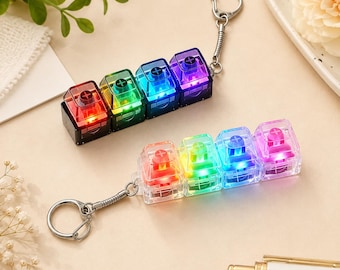 Puede incluir: Dos llaveros de interruptores de teclado mecánico, cada uno con cuatro teclas cuadradas y translúcidas en fila. El llavero superior tiene bases negras, mientras que el inferior tiene bases transparentes. Ambos emiten luz LED multicolor.