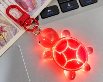 May include: A bright red, translucent turtle-shaped keychain featuring a glowing light effect. The turtle has a smooth, rounded shell with white line detailing and a matching red metal lobster clasp attached by a small silver ring.
