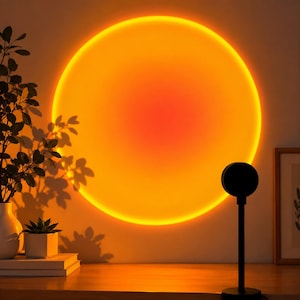 Könnte beinhalten: Eine Sonnenuntergangs-Projektionslampe mit einem schwarzen runden Kopf und einem dünnen Ständer, die ein großes, leuchtend orangefarbenes und gelbes kreisförmiges Licht an die Wand wirft. Das Licht erzeugt einen warmen Schein im Raum.