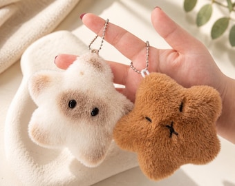May include: Two plush star-shaped keychains with metal ball chains. One star is cream-colored with brown patches around two black eyes. The other star is solid brown with a stitched black "x" for a mouth and two small horizontal lines for eyes.