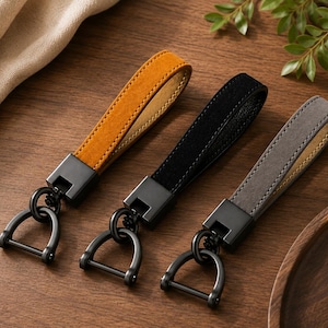 May include: Three keychains featuring looped straps in mustard yellow, black, and grey suede-like material. Each strap is attached to a matte black metal square connector and a matching black metal D-shaped shackle with a screw-pin closure.