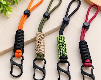 May include: Four paracord lanyards with black carabiner clips. Each lanyard features a loop with a black plastic cord lock and a braided section. The colors include orange and black, olive green and tan, black and neon green, and maroon and black.