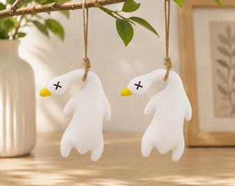 May include: Two white goose-shaped ornaments hanging from a branch by brown twine. Each goose has a bright yellow beak and black 'X' marks for eyes. The figures have a smooth, matte finish and are depicted in a downward-facing, hanging position.