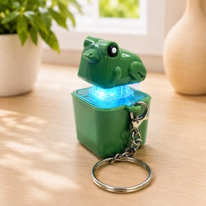 May include: A green frog-shaped keychain featuring a square base and a top section that glows with a bright blue light. The object includes a silver-toned metal split ring attached by a small chain, allowing it to be used as a functional accessory.