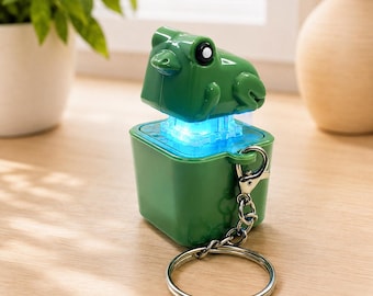 May include: A green frog-shaped keychain featuring a square base and a top section that glows with a bright blue light. The object includes a silver-toned metal split ring attached by a small chain, allowing it to be used as a functional accessory.