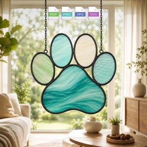 May include: A stained glass suncatcher shaped like a paw print, featuring five individual segments with marbled textures in shades of teal, light green, and cream, all encased in a black metal frame and suspended by two thin black metal chains.
